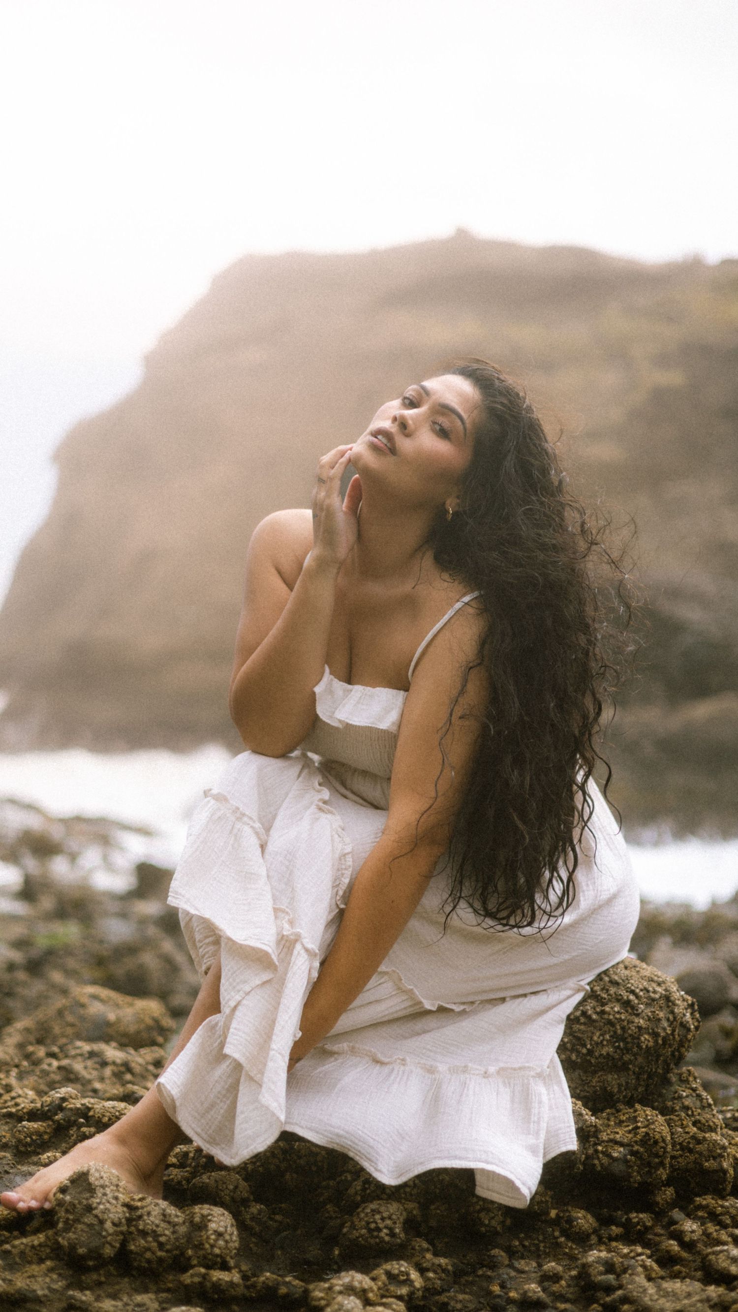 A person in a flowing white dress poses dramatically on coastal rocks during golden hour at the beach.