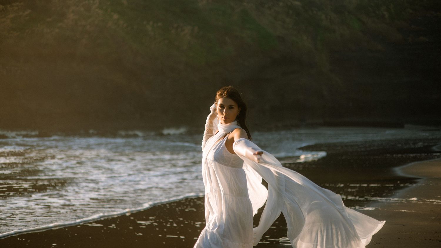 Flowing white dress creates artistic movement against dark volcanic beach and ocean backdrop.