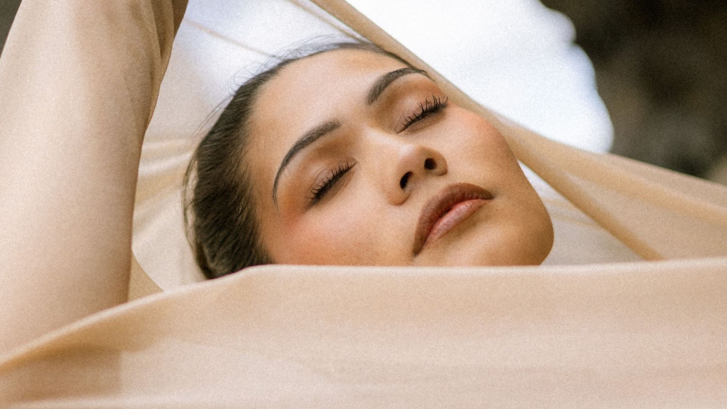 A serene woman with closed eyes rests peacefully against soft beige fabric in natural light.