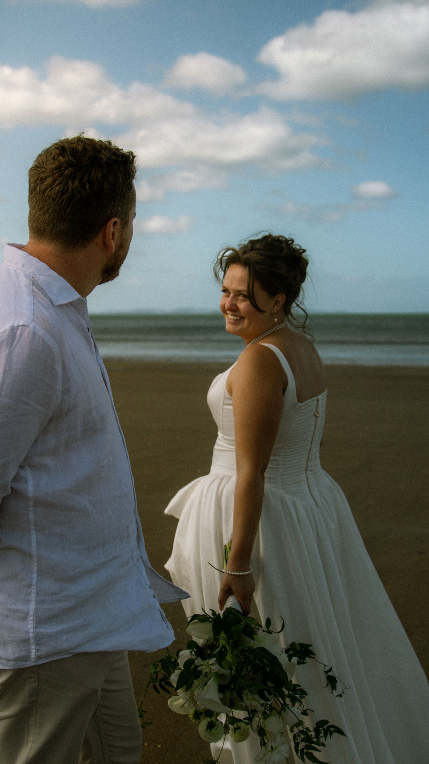 A smiling bride in a white gown holds a bouquet while standing with her groom on a sandy beach.