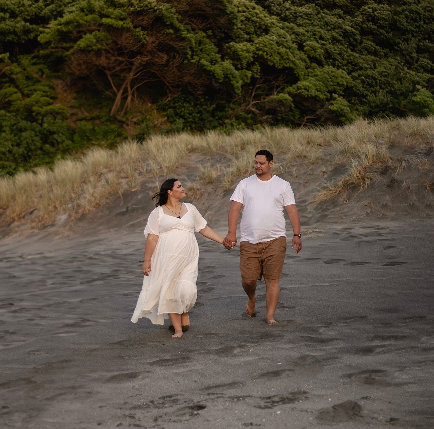 Two people walk together on a dark sandy beach with lush green trees in the background during golden hour.