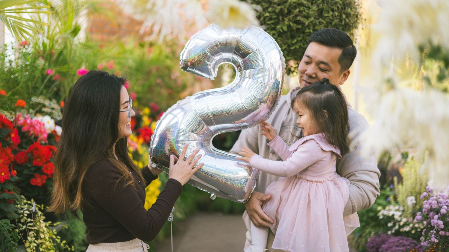 Family celebrating a 2nd birthday with a silver balloon number 2 among colorful flowers.