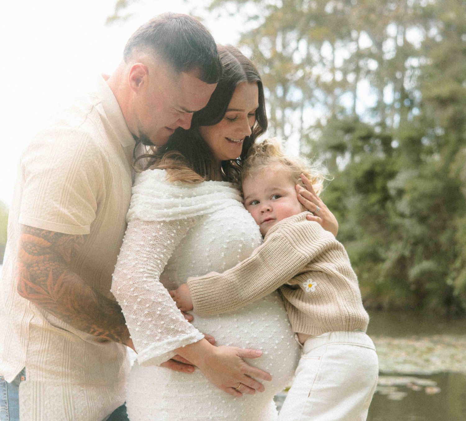 Expecting couple embracing a pregnant belly while a toddler hugs mom, in a soft outdoor maternity photo beside a tranquil lake.
