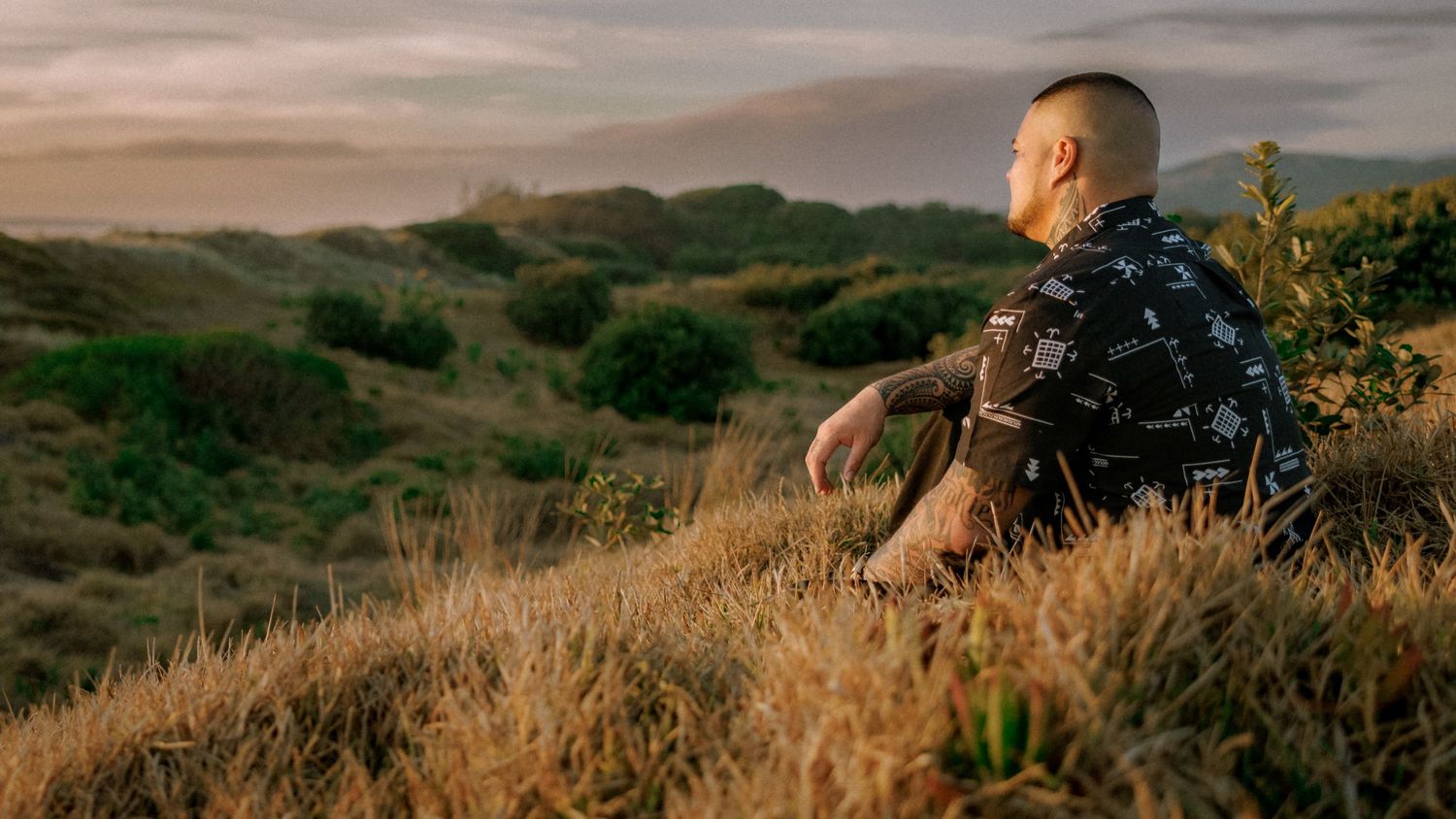 Person in black shirt sits on grassy hillside during golden hour overlooking scenic landscape at sunset.