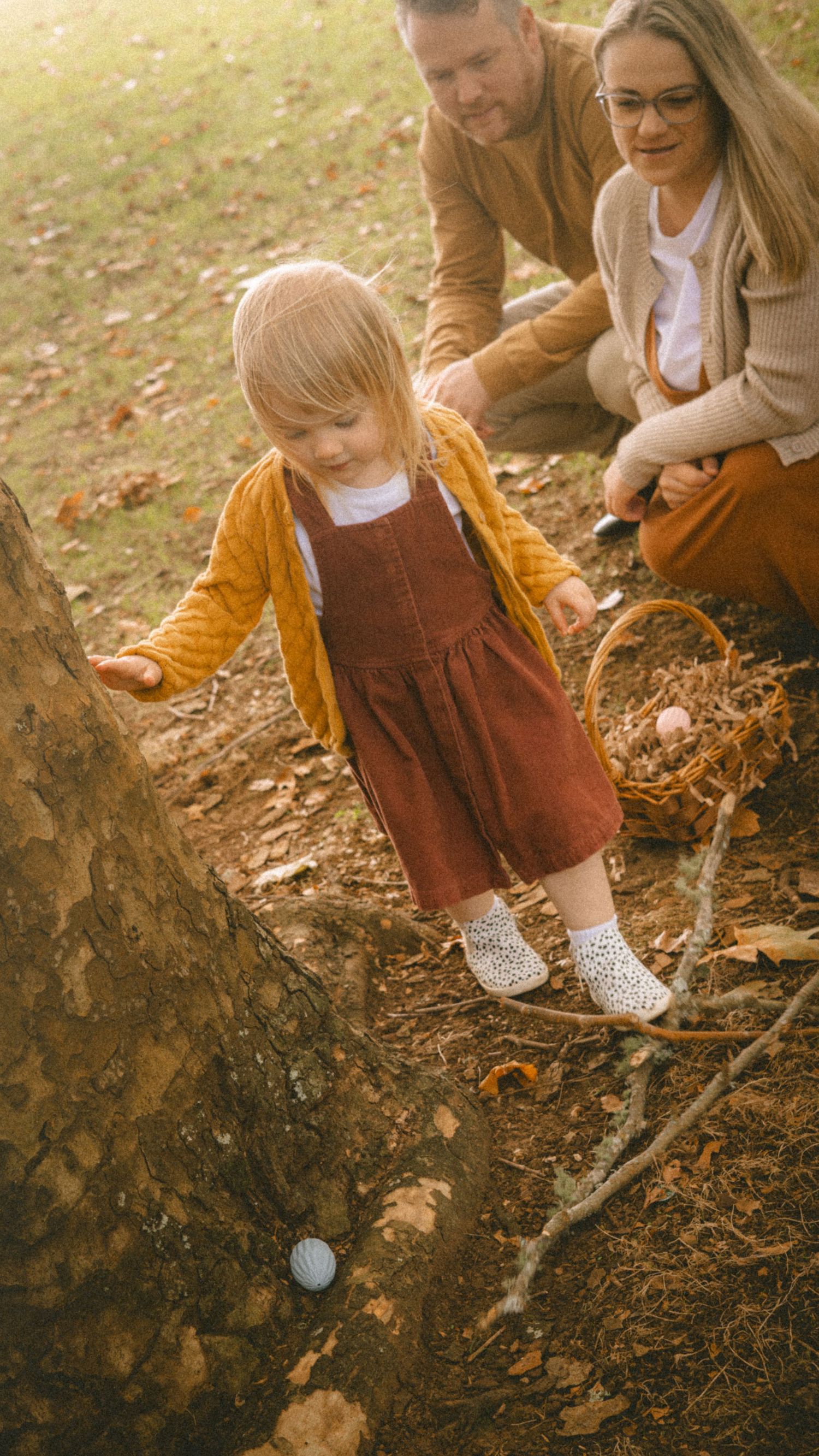 A toddler girl in a red dress discovers an Easter egg near a tree while her parents watch during an outdoor egg hunt.