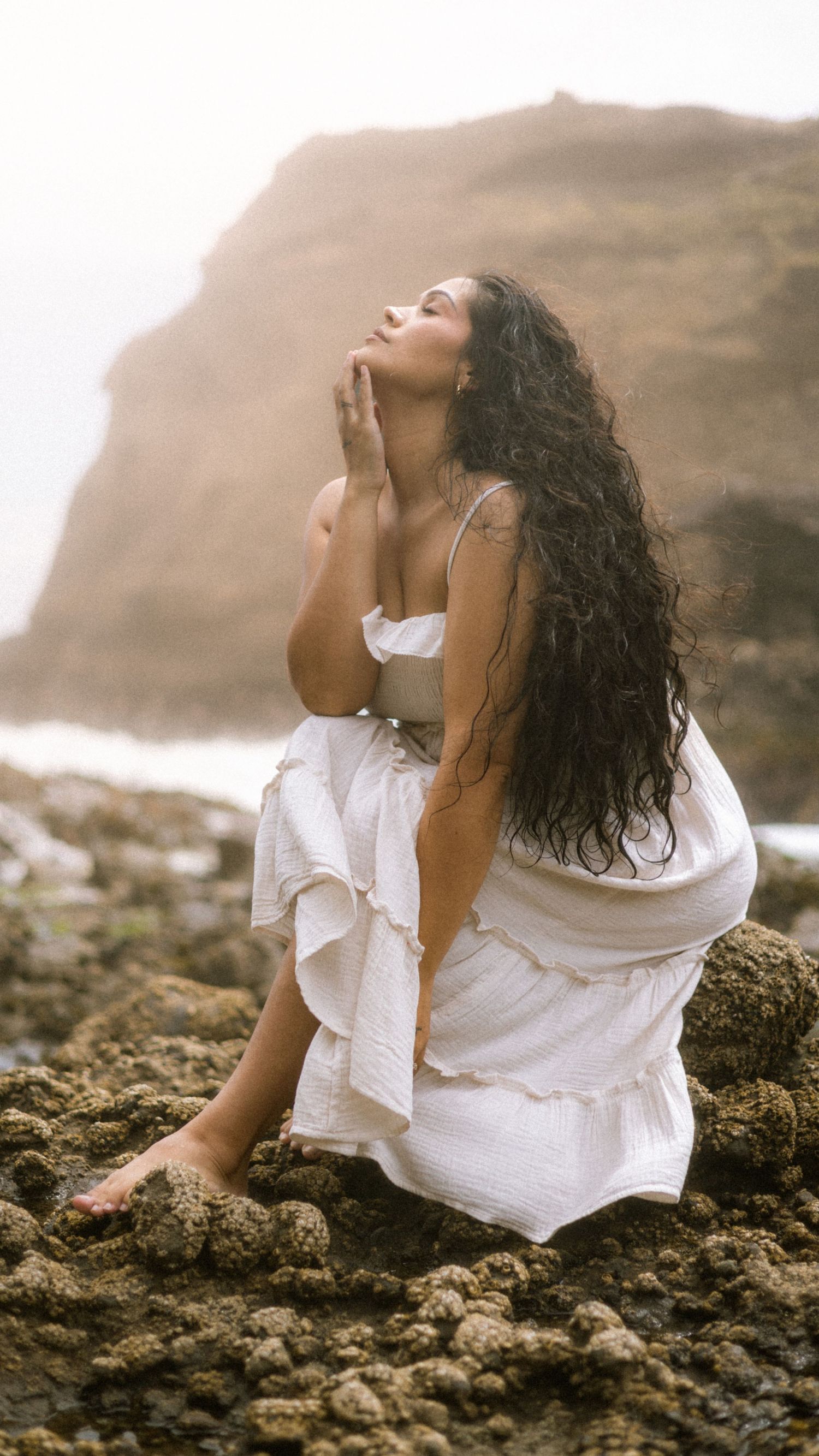 A person in a flowing white dress poses gracefully on rocky coastal cliffs with long dark wavy hair and misty backdrop.