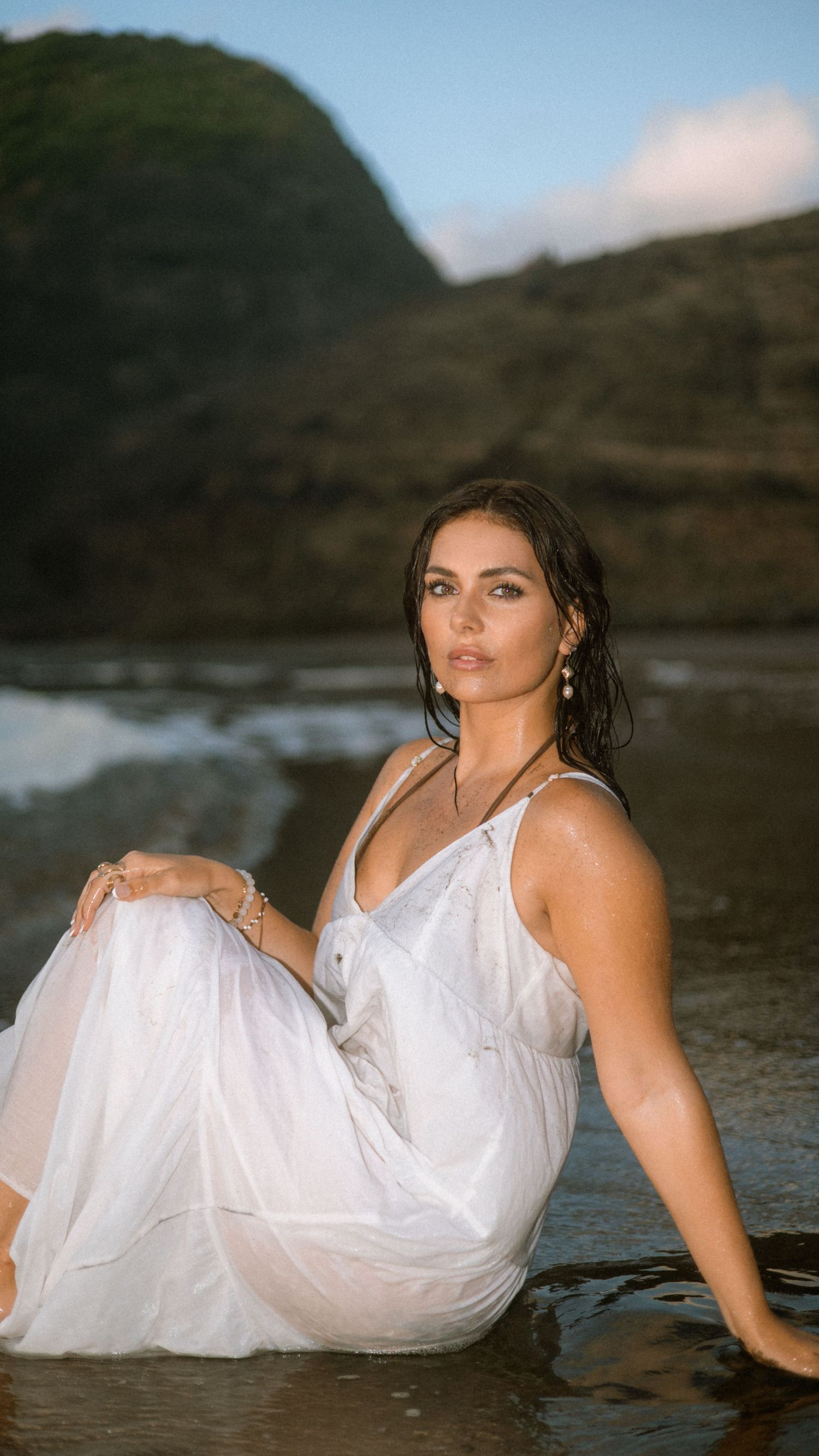 Model in flowing white dress poses gracefully by the water's edge against a mountainous backdrop.