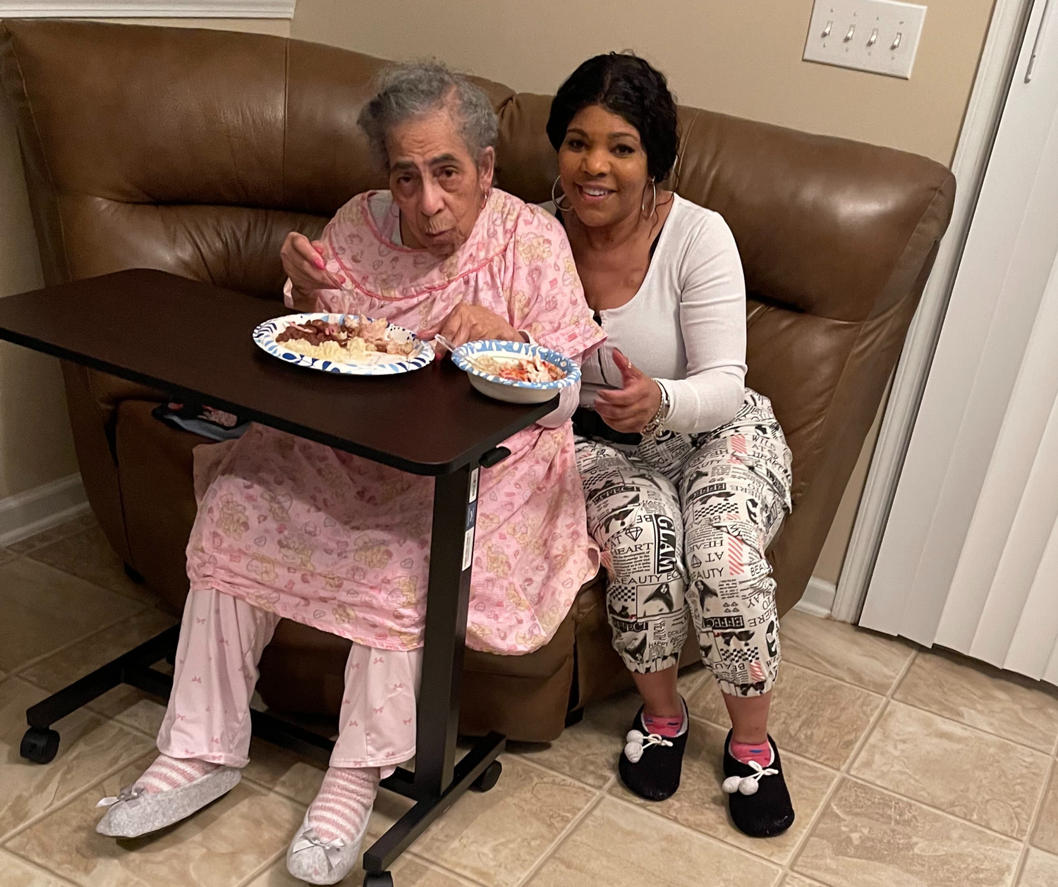 Thrive Living Services Employee with client sitting together on a brown leather couch. Providing a meal from a portable tray table.
