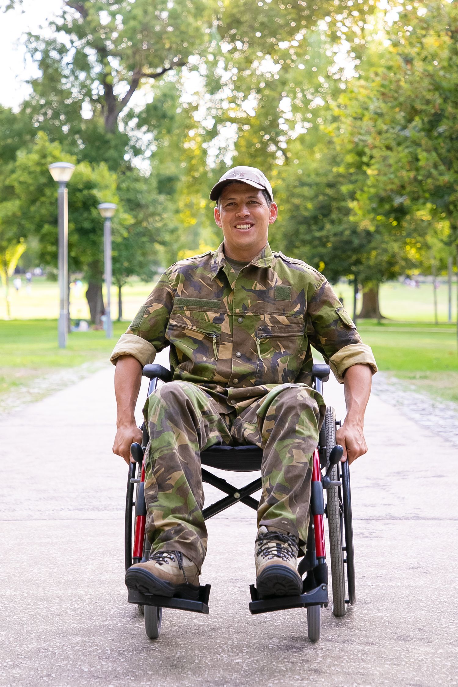 Military service member in camouflage uniform sits in wheelchair on park path with trees and lamp post in background.