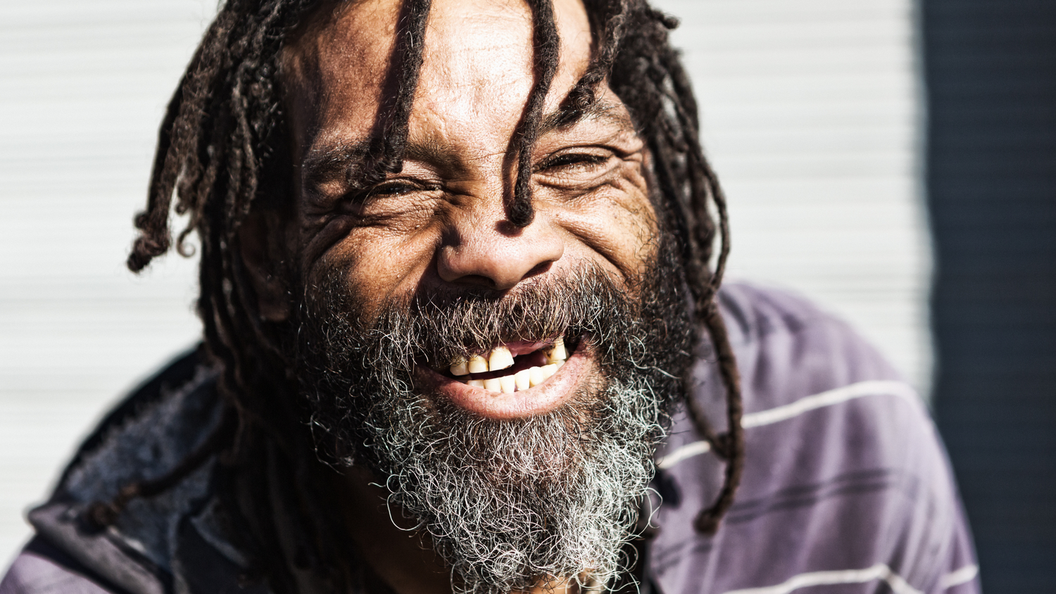 A bearded homeless man with dreadlocks laughs joyfully while wearing a purple shirt in portrait.