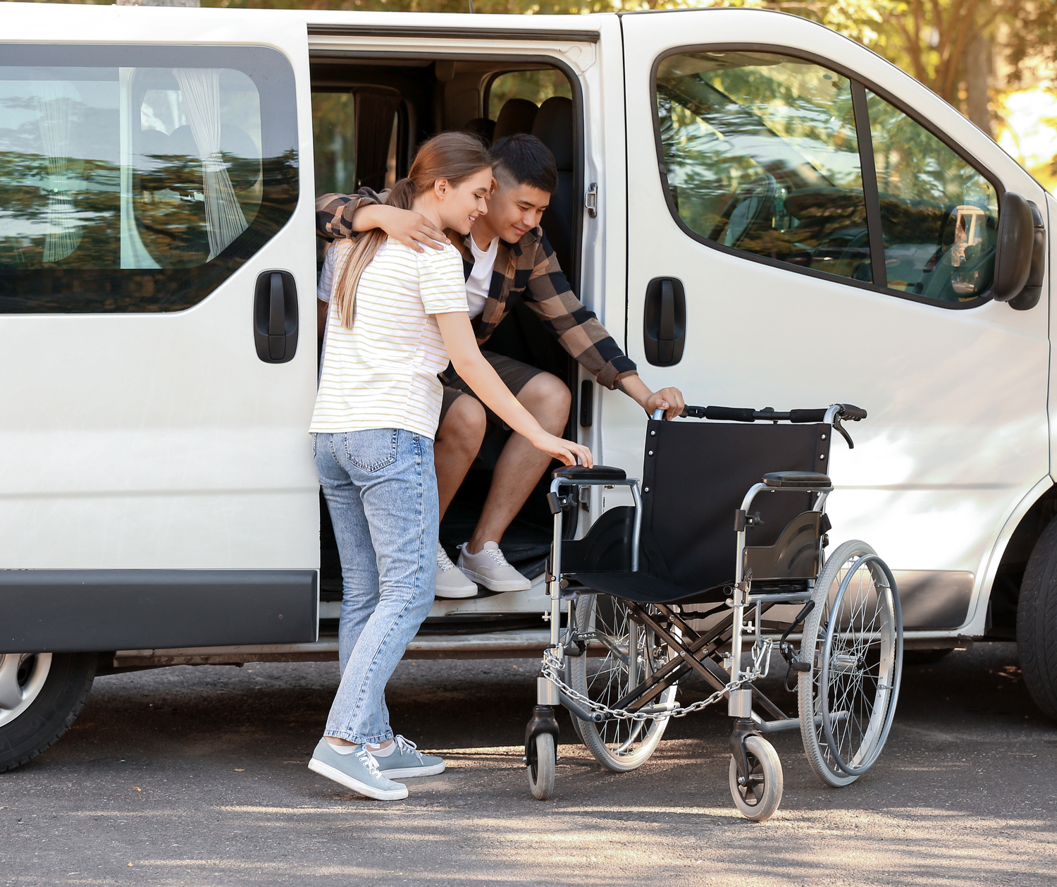 A caregiver assists someone getting into a wheelchair from a white passenger van in a parking area.