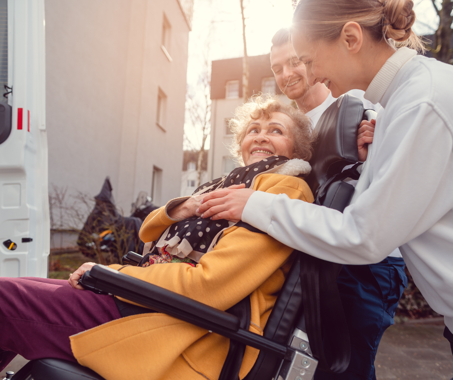 A person in a yellow jacket sits in a wheelchair while another person assists with their care in a medical setting.
