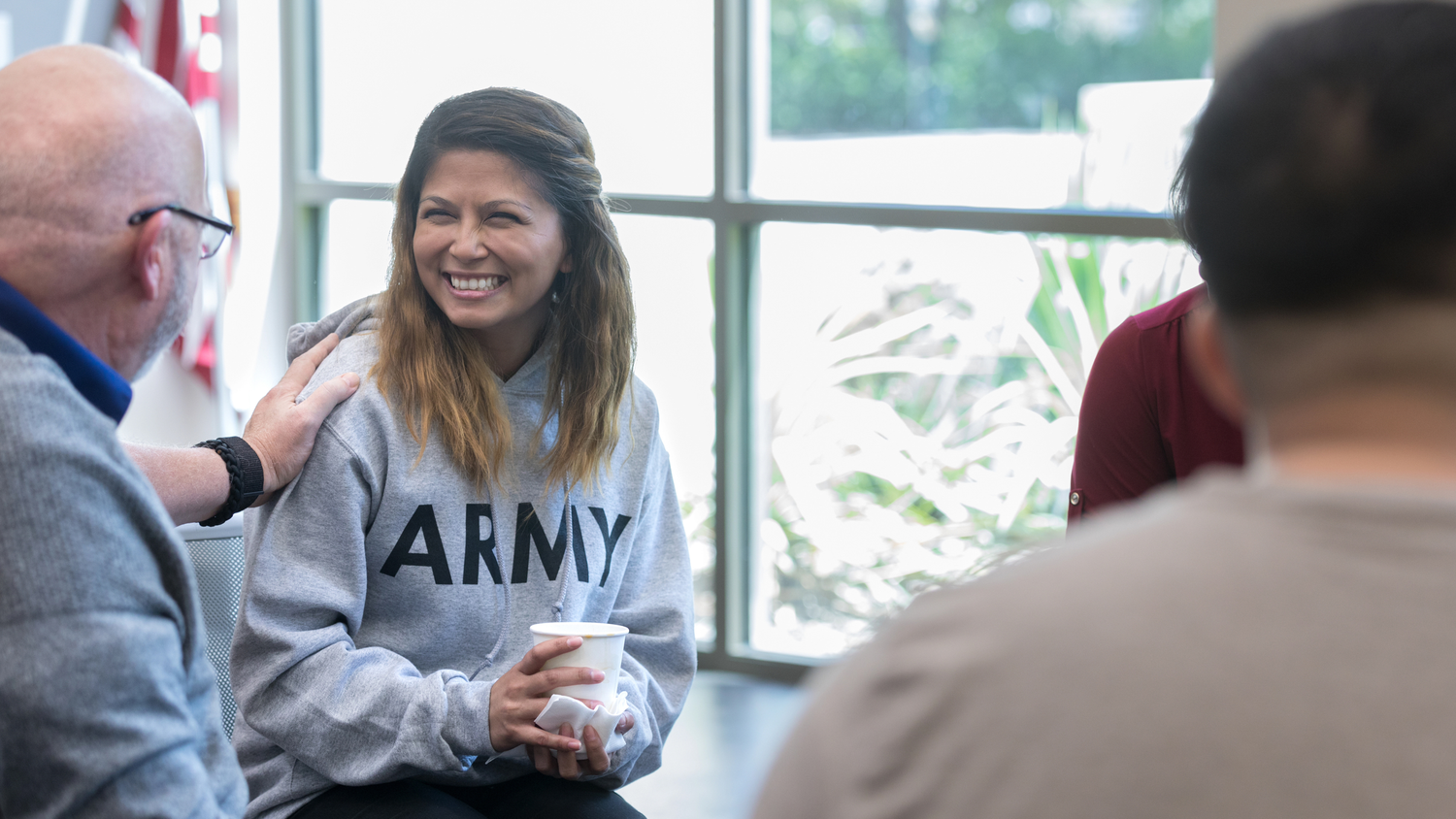 A group of people and army vet sitting at tables in a bright office space having casual conversations during a meeting.