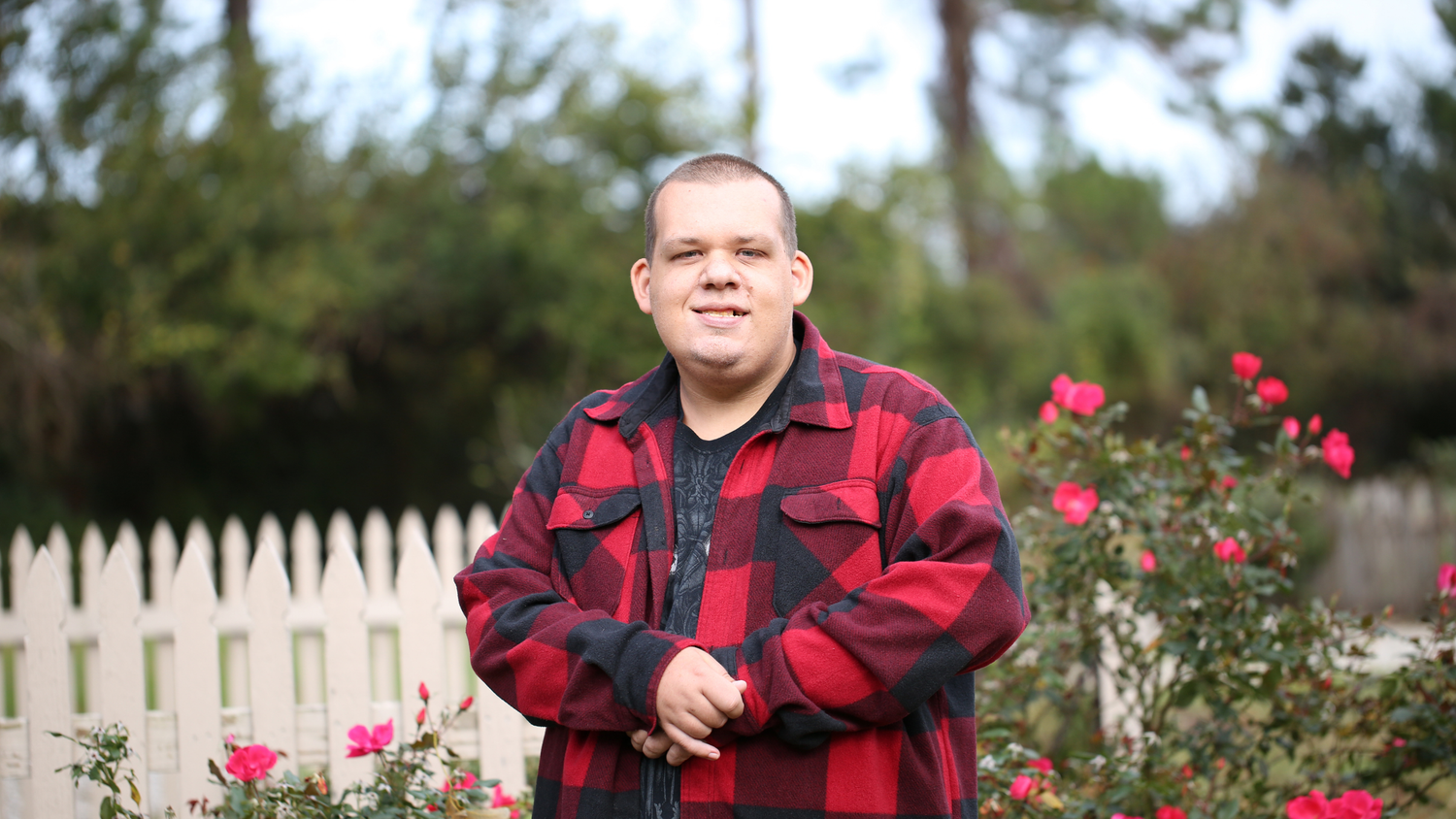A person wearing a red and black buffalo plaid flannel shirt poses outdoors near a white picket fence and garden.