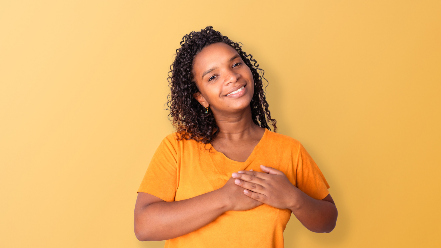 A lady standing against an orange background showing and gesturing love. With hands over chest.