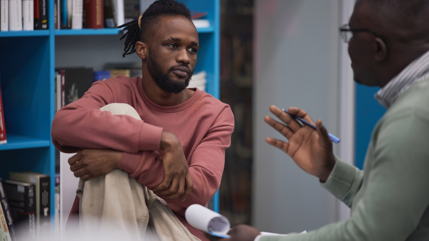 Black male in n pink sweater gestures expressively during animated conversation in private therapy session.