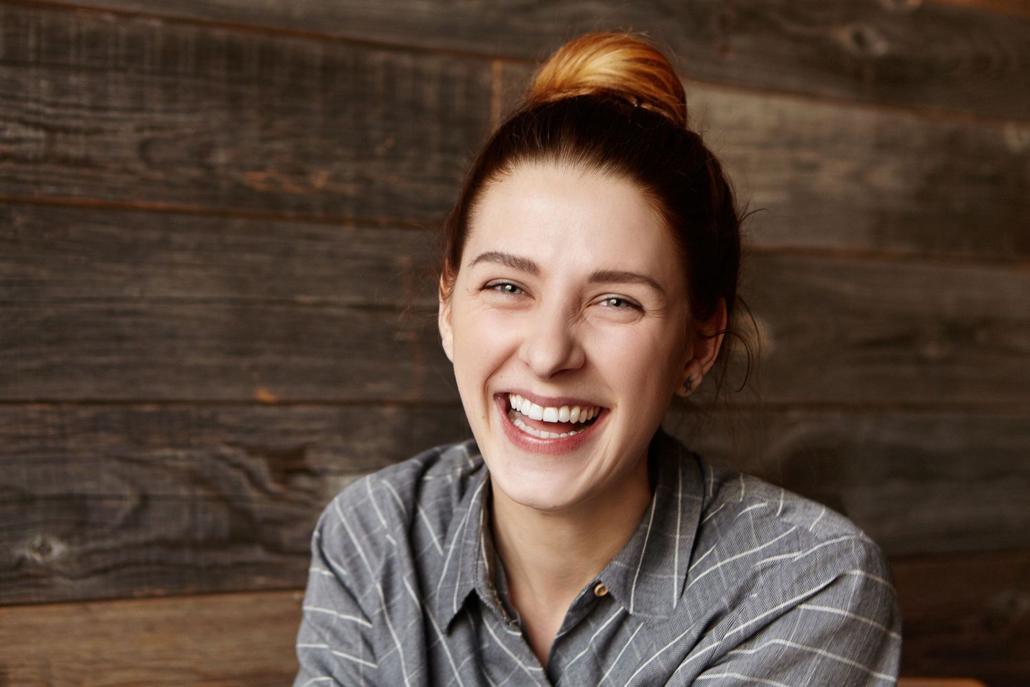 A smiling person wearing a gray sweater poses against a rustic wooden wall background.