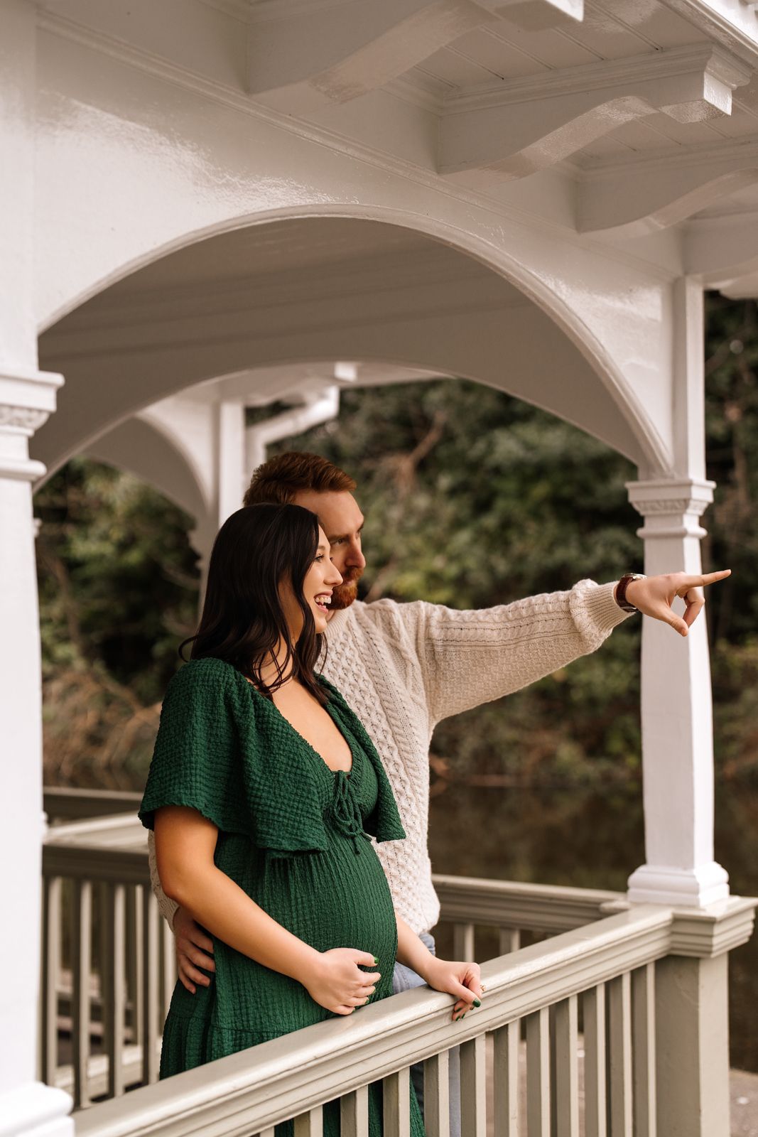 A pregnant woman stands on a porch, pointing and looking into the distance.