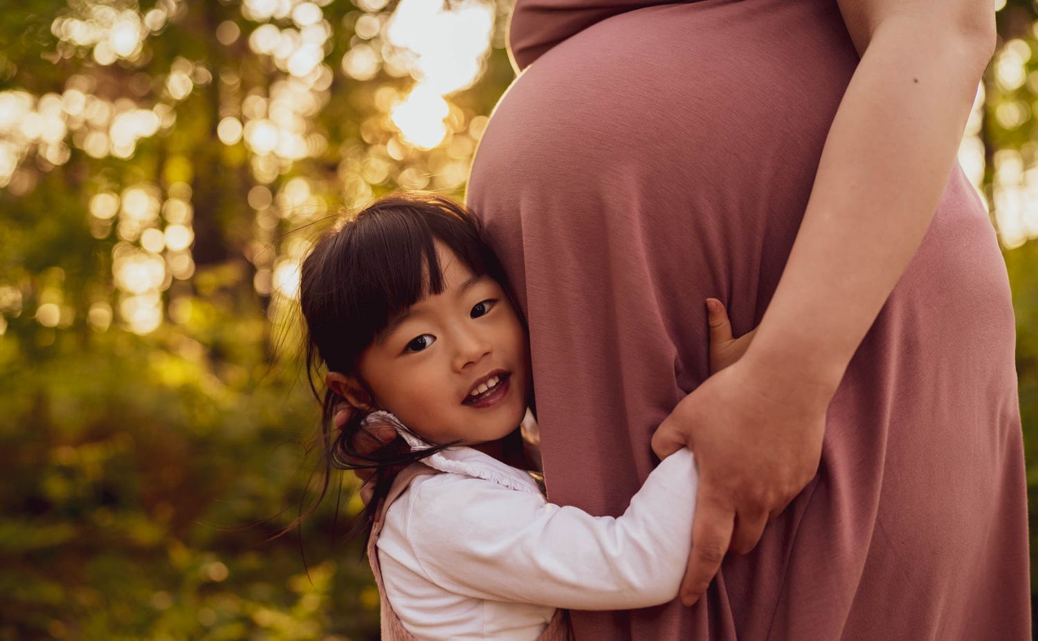 A small child hugs a pregnant woman's belly in a sunlit forest setting.