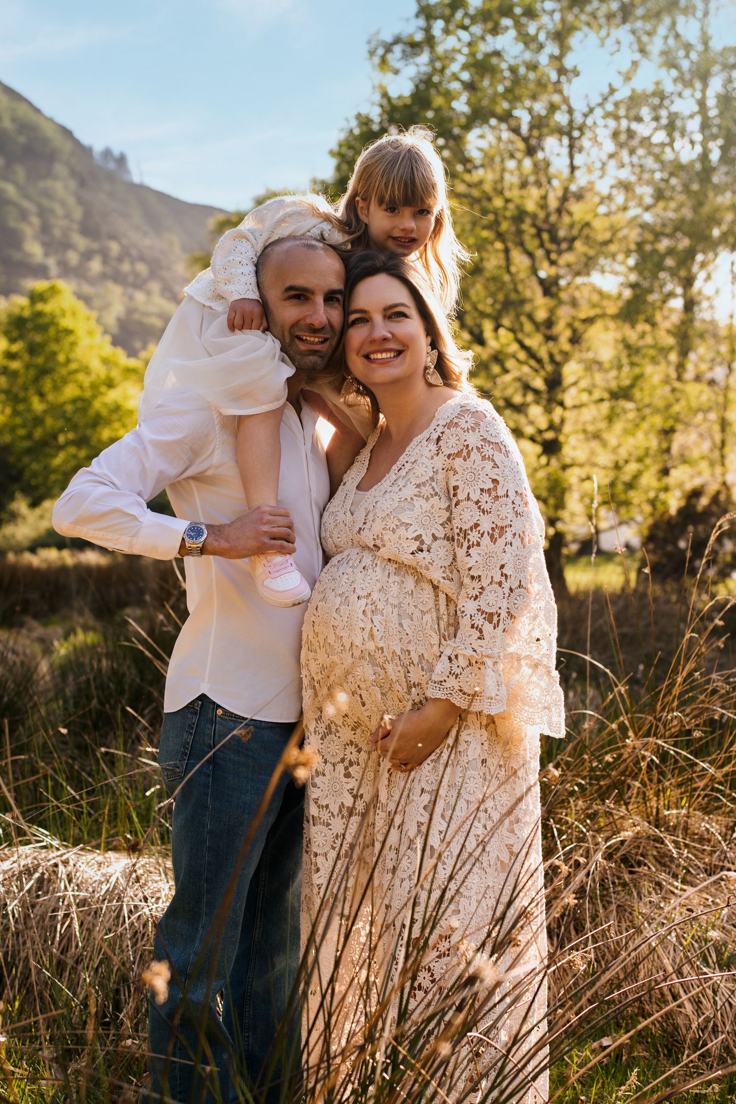 Couple embracing in field of tall grass with mountain backdrop during golden hour.