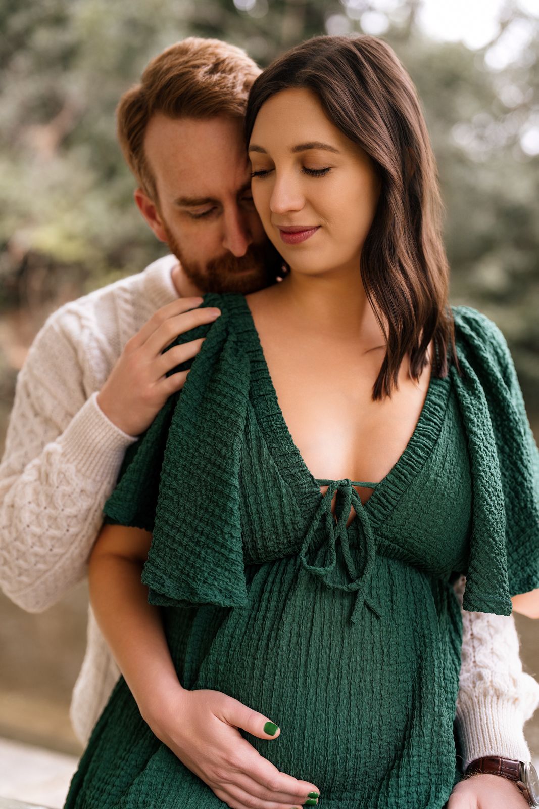 A pregnant woman in a green dress is embraced by her partner in a warm outdoor setting.