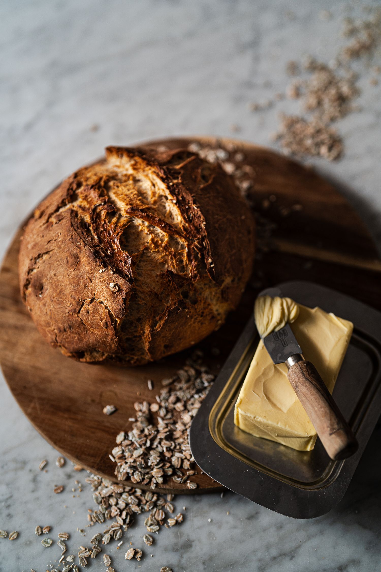 Fresh baked artisanal dark bread loaf on wooden board with butter dish and scattered seeds.