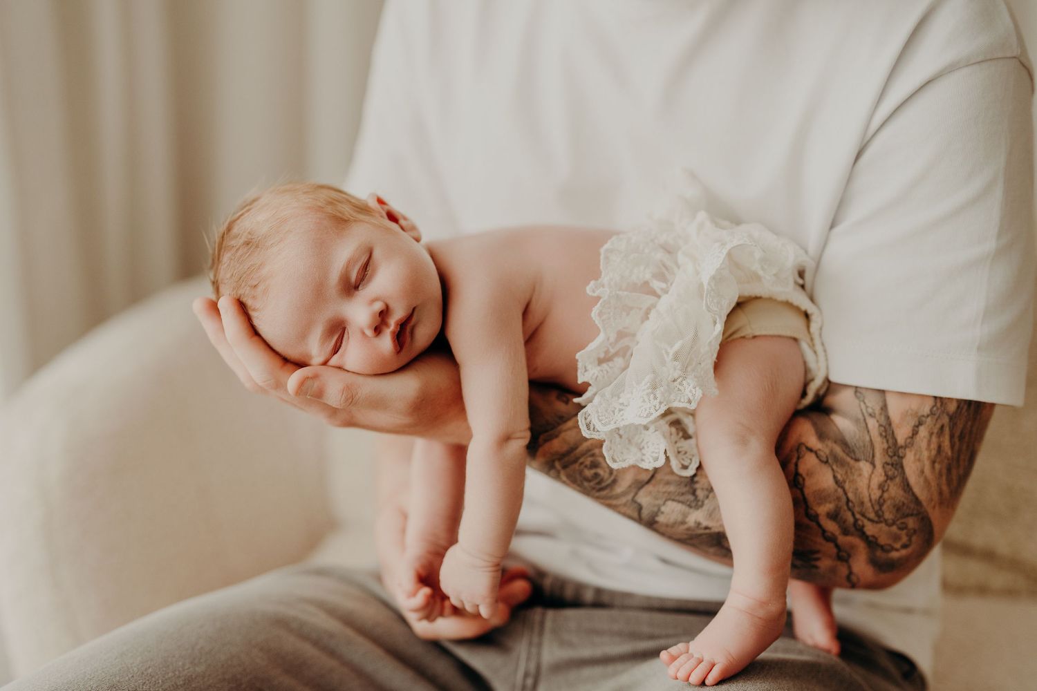 Sleeping baby wearing white lace clothing while resting on parent's lap.