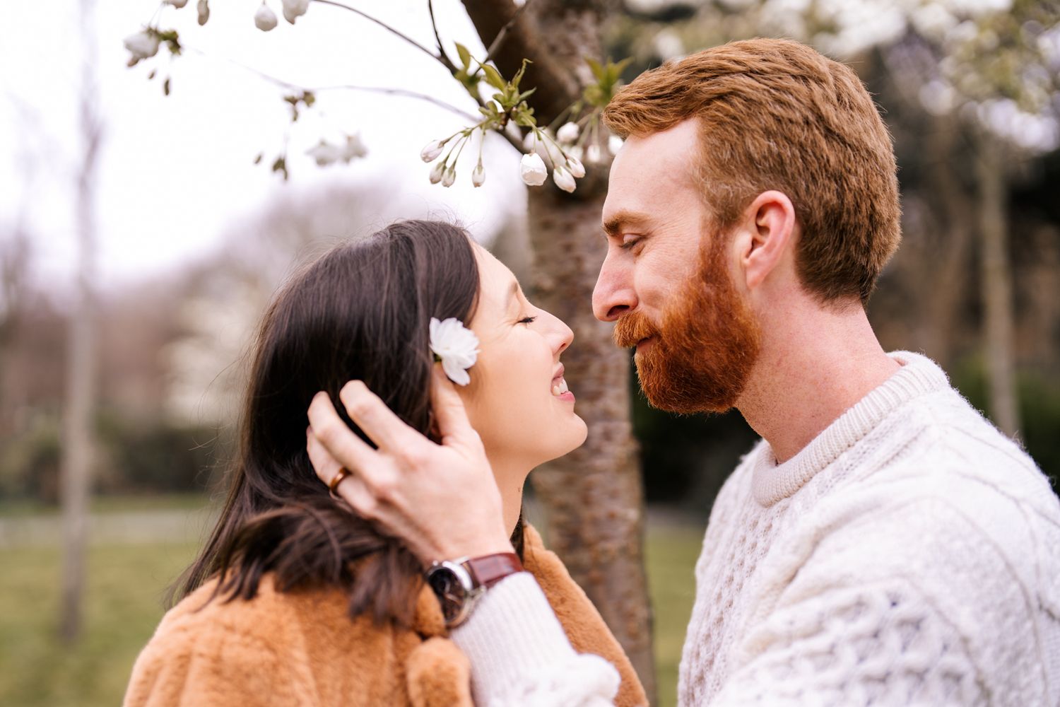 A man and woman embrace tenderly in a park setting with blooming trees in the background.
