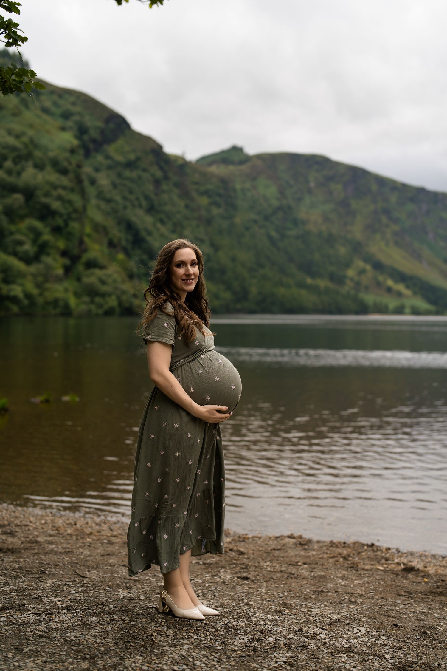 A woman in a polka dot dress sits by a serene lake surrounded by mountains on a cloudy day.