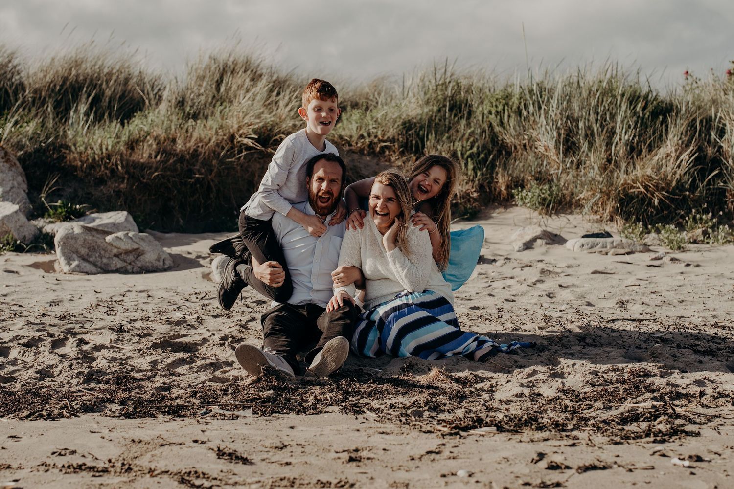 Candid family moment captured on a peaceful beach with tall grass in the background.