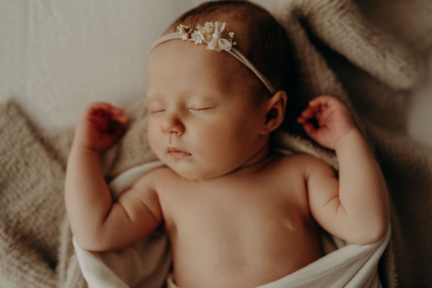 A sleeping newborn baby wearing a delicate floral headband rests peacefully on soft fabric.