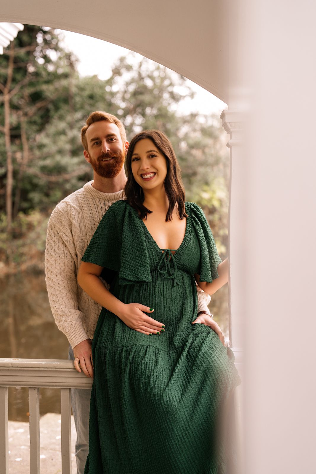 Couple posing on elegant white balcony, woman in green maternity dress cradling baby bump, man in cream sweater behind her.