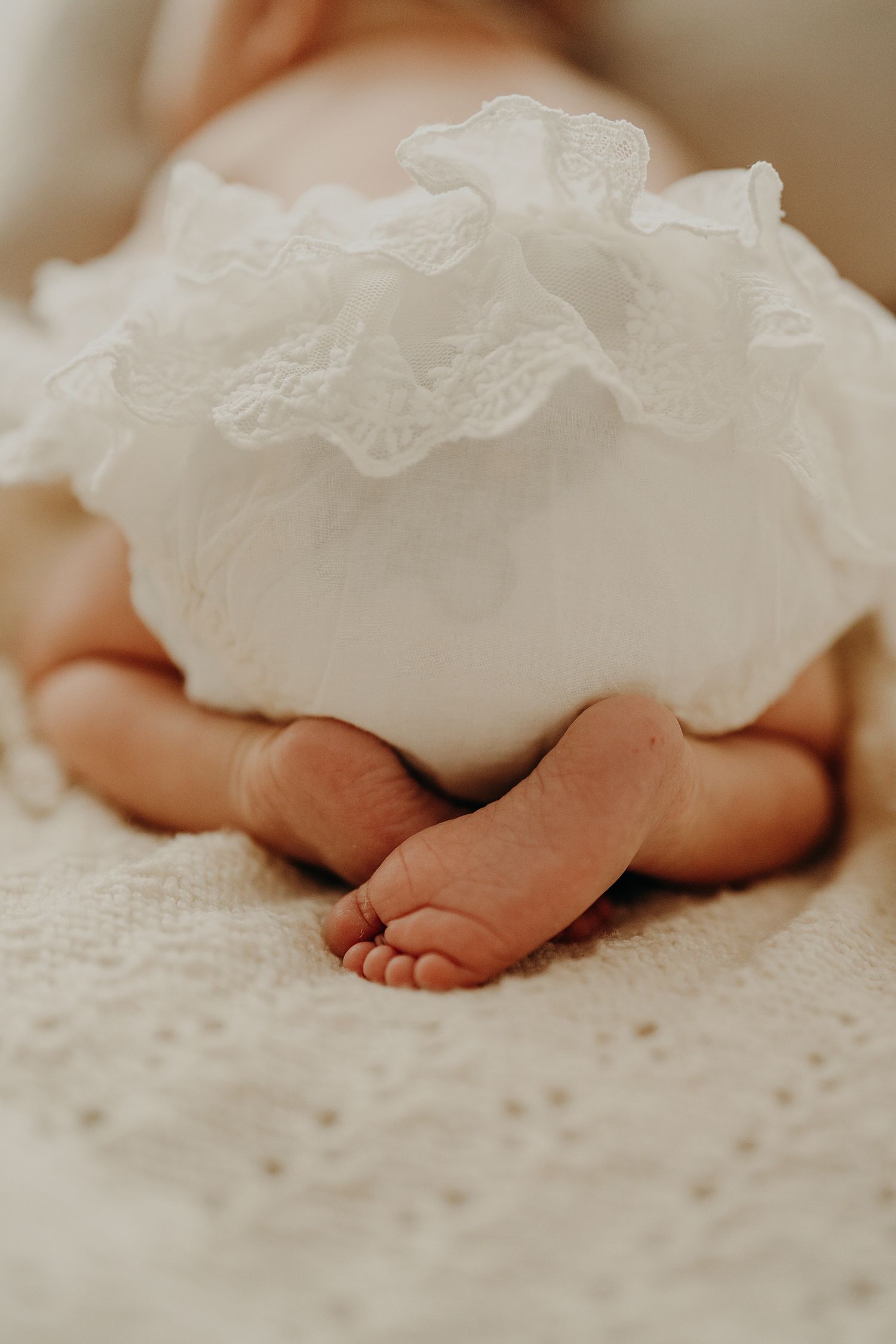 Tiny baby feet peeking out from under white lace dress against textured fabric.