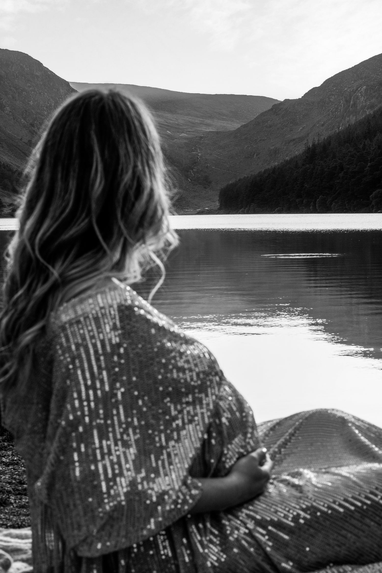 Person in sequined outfit photographed in black and white against scenic mountain lake backdrop.