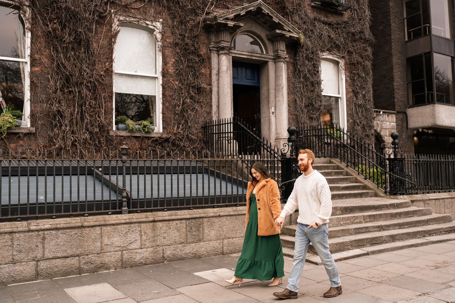 A couple walking together in front of a historic building with ivy-covered walls.