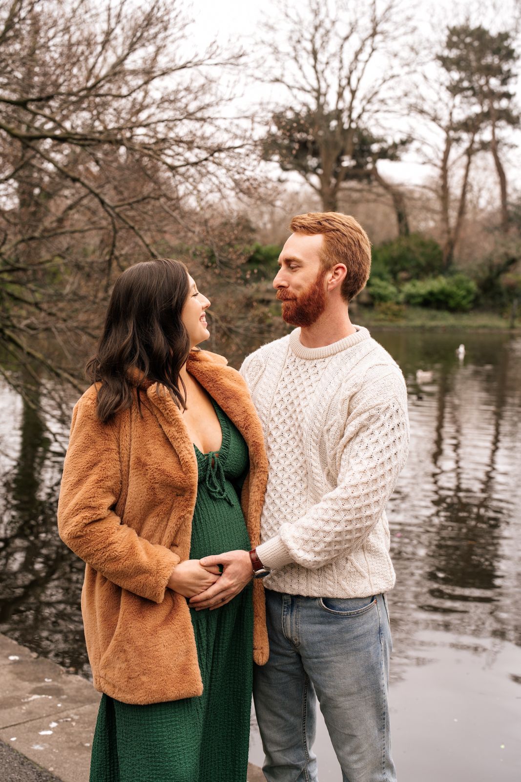 A couple stands together by a pond, surrounded by trees and nature, creating a serene and romantic scene.
