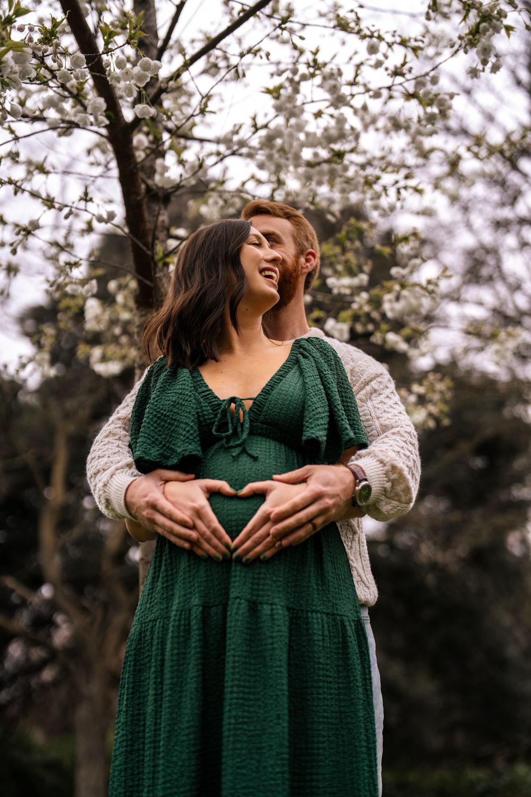 Couple embracing outdoors, man forming heart shape on pregnant woman's belly in green dress near blooming tree.
