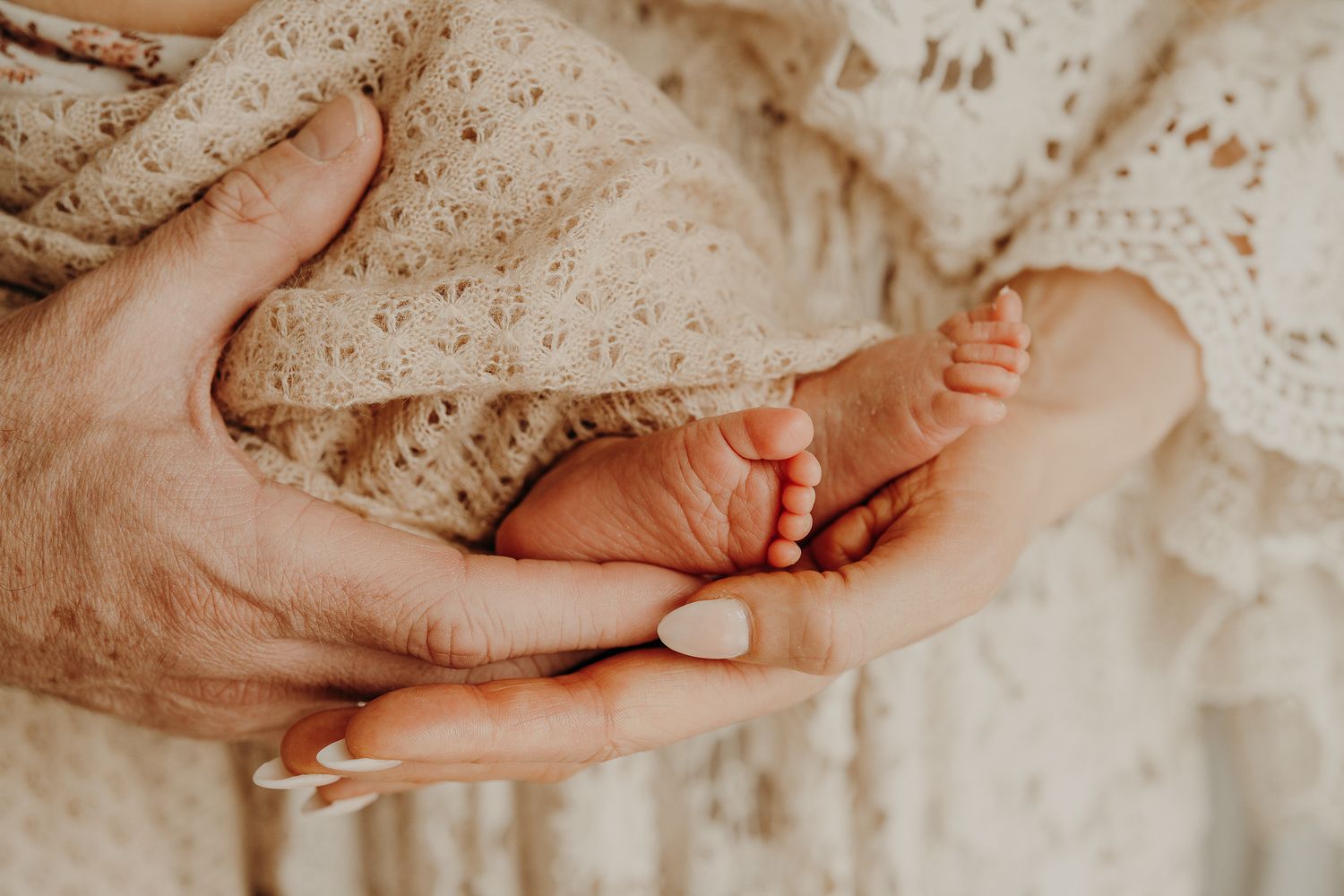 Close-up of tiny baby feet being gently held in adult hands against lace fabric.