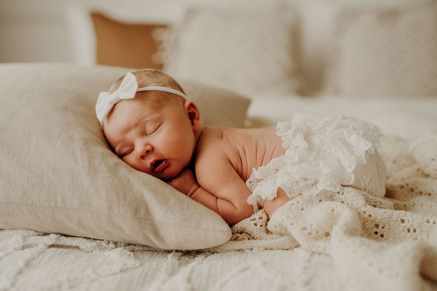 Sleeping newborn with white bow headband resting peacefully on light colored pillows.