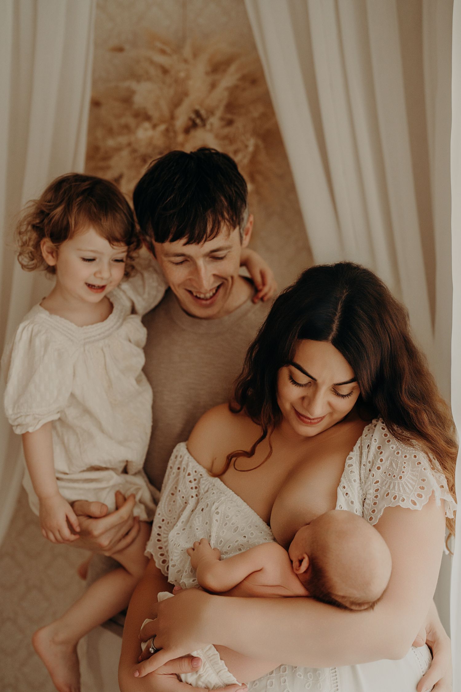 Intimate family moment with nursing baby and toddler in white curtained setting.