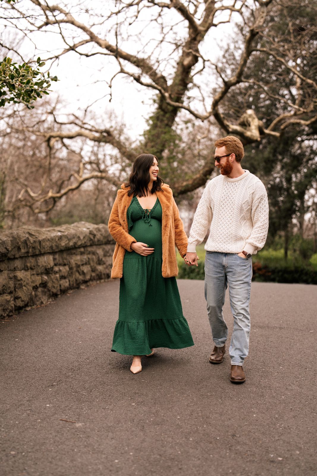 A pregnant woman in a green dress and a man in a white sweater walk together on a path surrounded by trees.