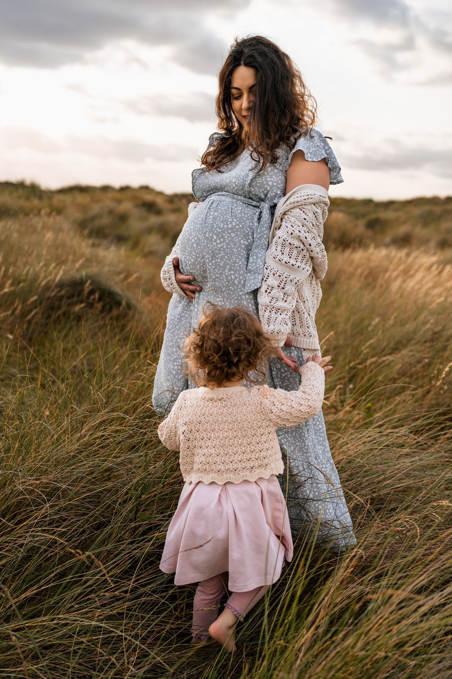 A pregnant woman in a gray dress stands with a small child in a grassy field.