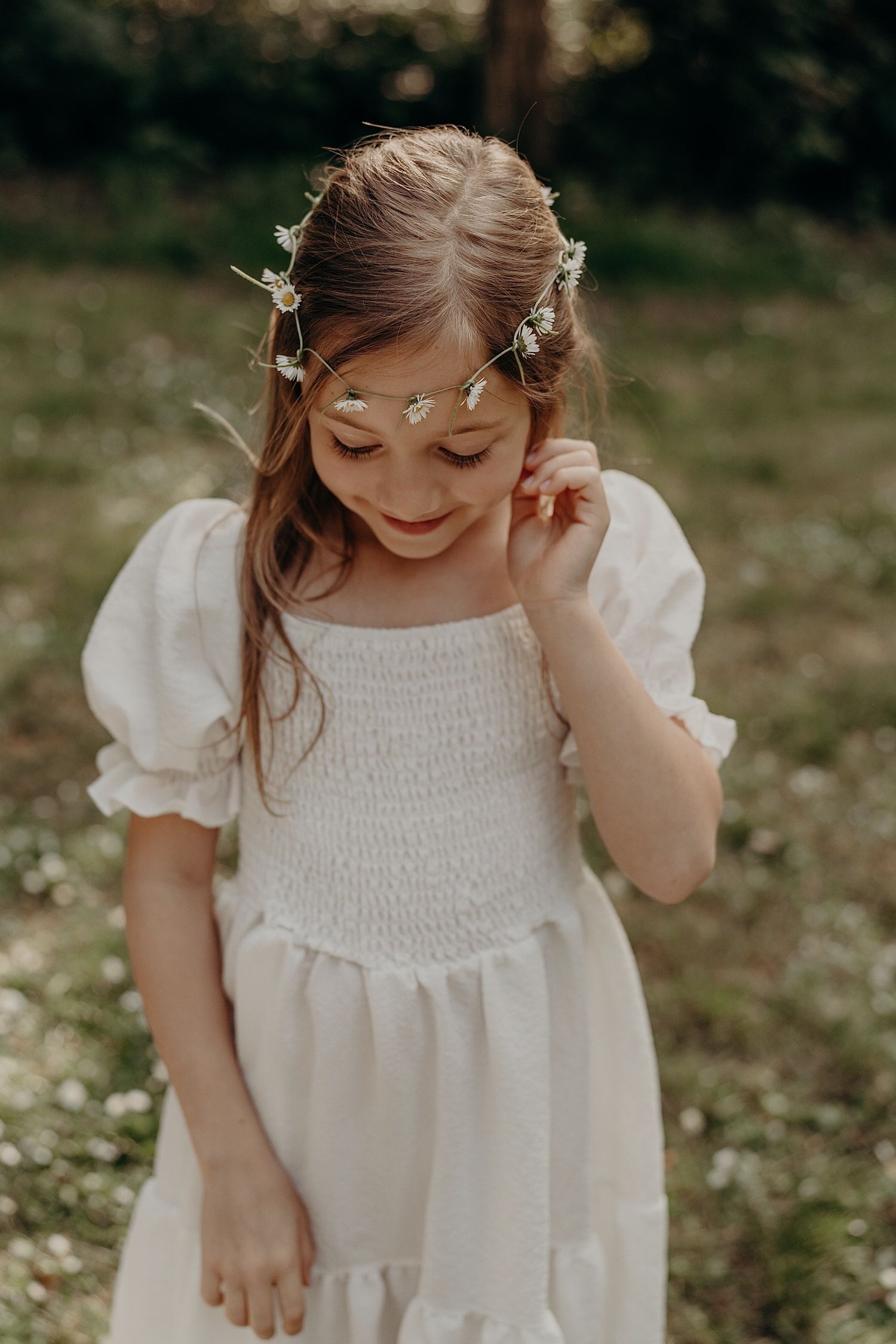 Young person in white smocked dress smiling while adjusting hair with flowers outdoors.
