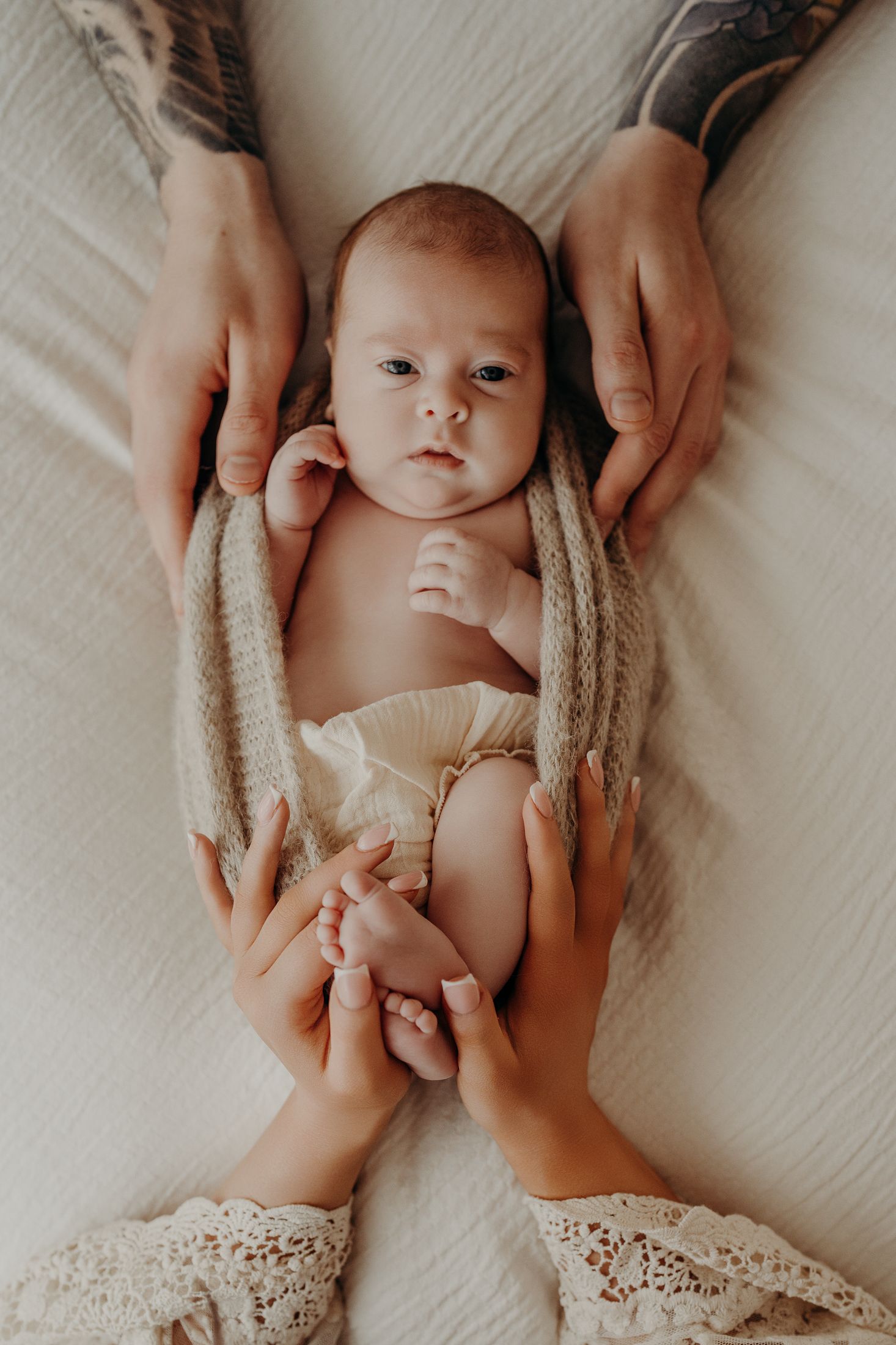 Overhead view of a baby lying on white bedding with hands gently cradling them from both sides. Natural newborn session Dublin