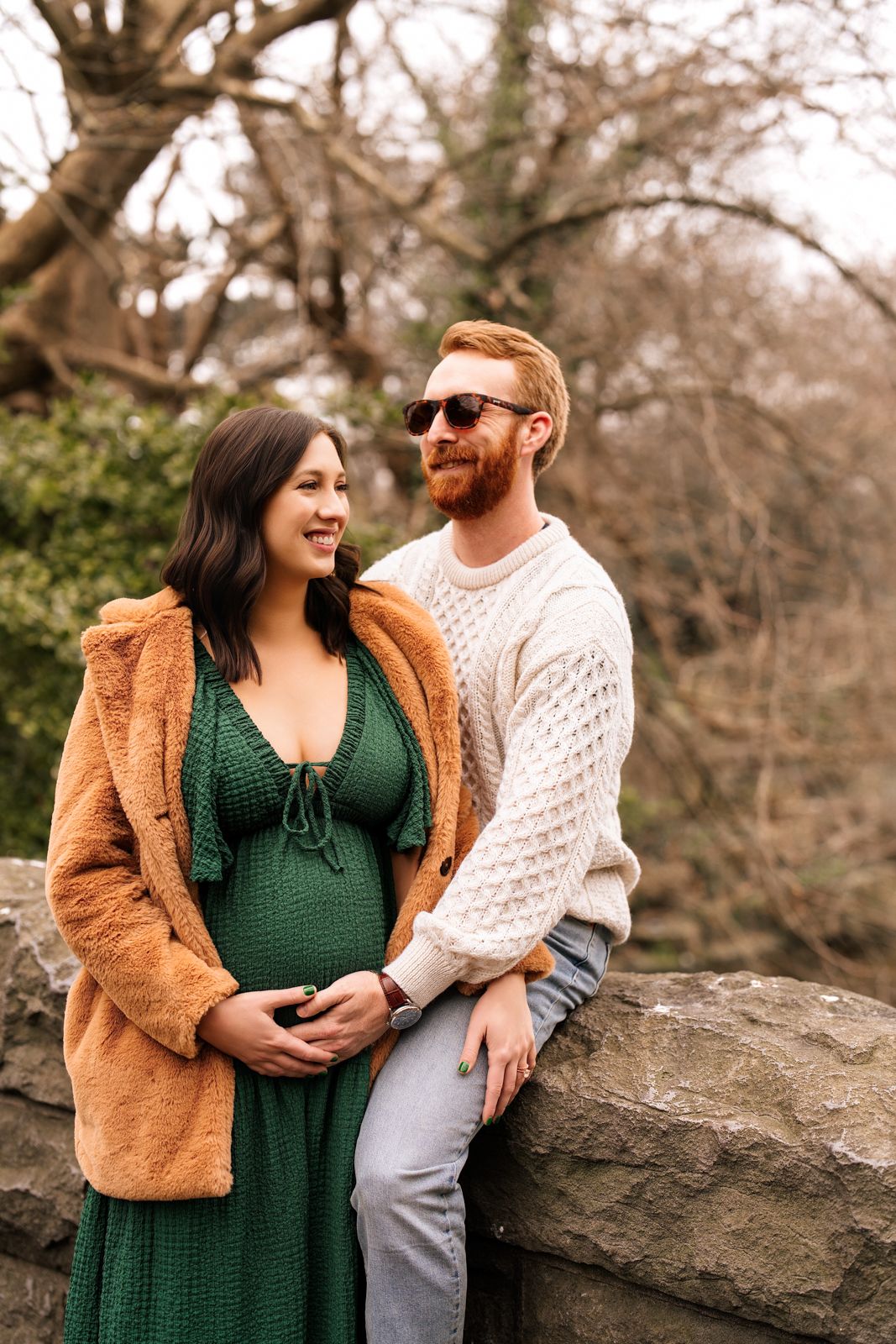 A pregnant woman in a green dress stands with her husband in a wooded area, smiling and embracing.