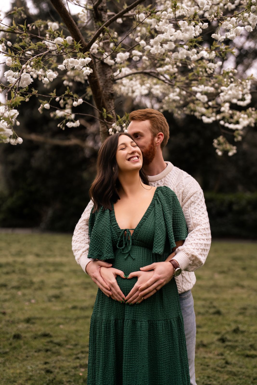 Couple sharing a tender moment under white blossoming tree, woman in green dress with partner forming heart shape on baby bump.
