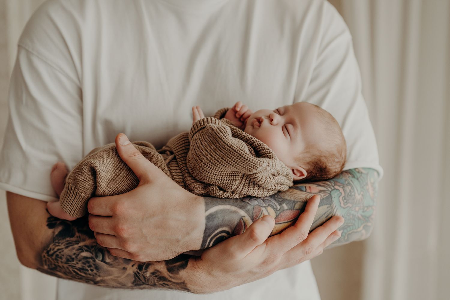 Gentle hands cradle a sleeping newborn baby wrapped in a soft knit blanket against a neutral backdrop.