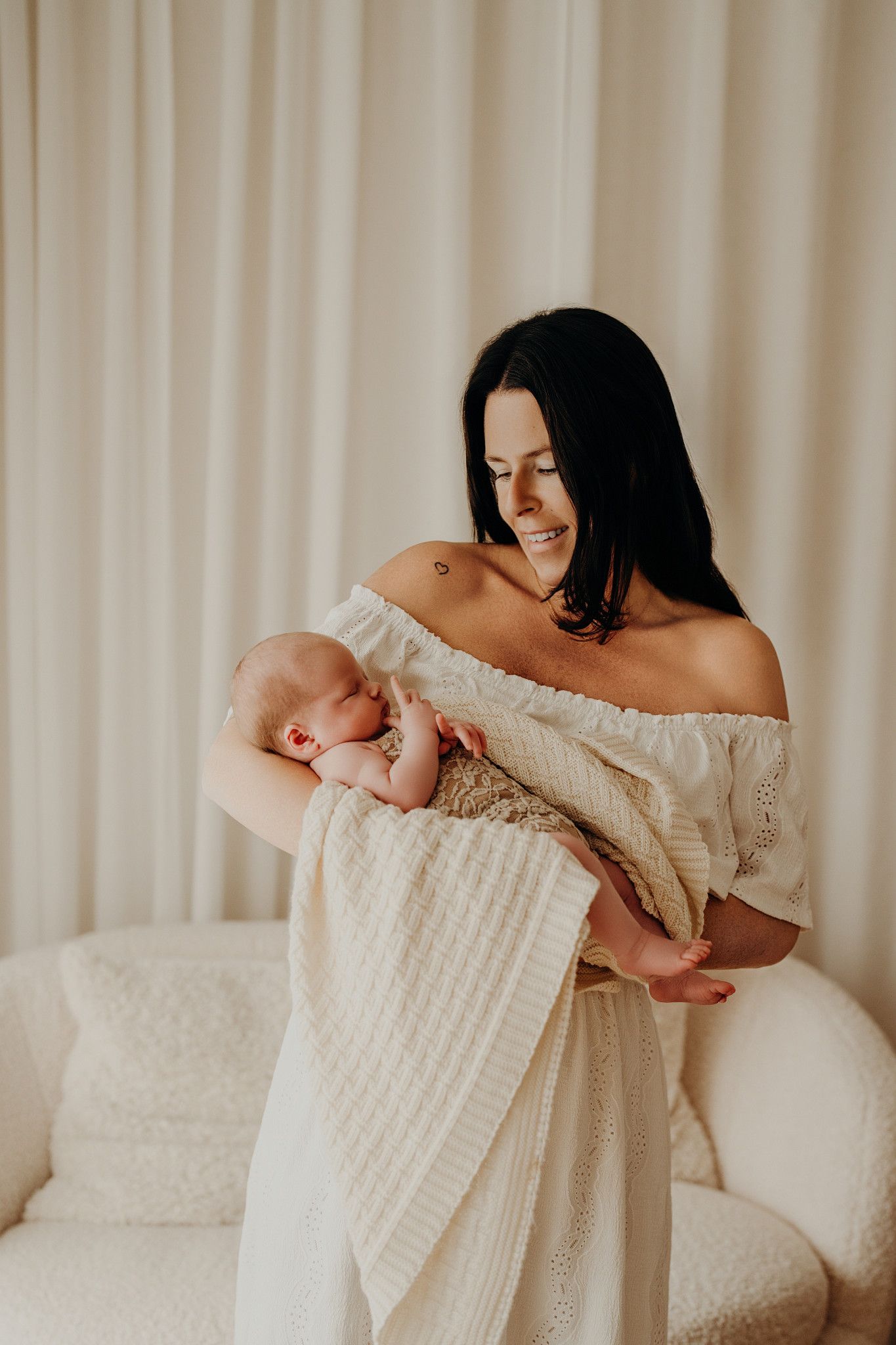 Mother embracing newborn baby wrapped in white knit blanket by cream curtains.