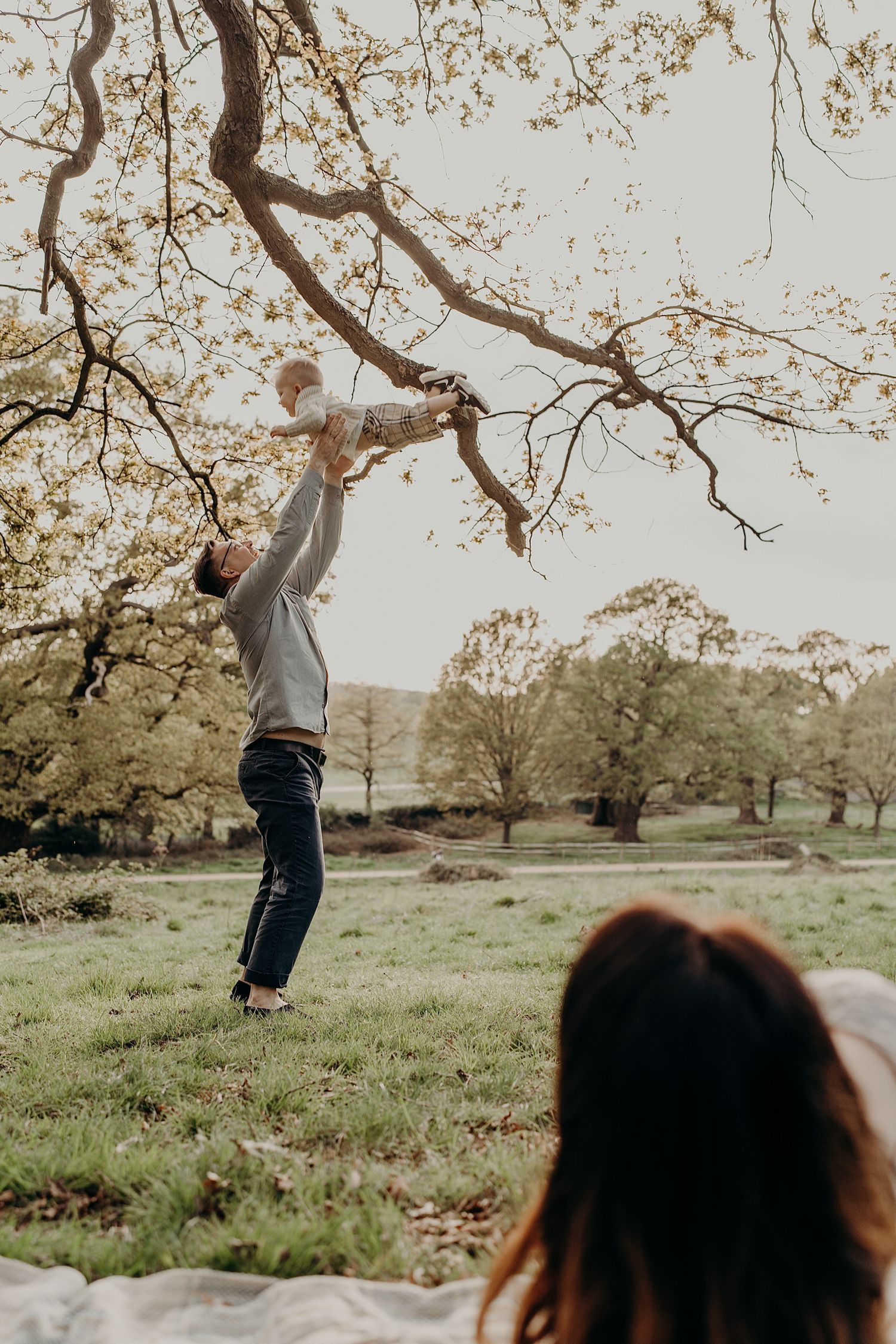 Child playing and swinging from tree branch in park while parent watches.
