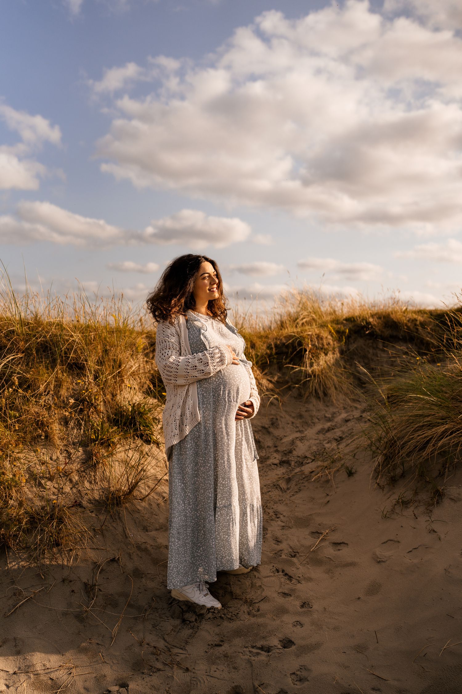 Woman in white lace dress poses on sandy dunes at sunset with dramatic cloudy sky.